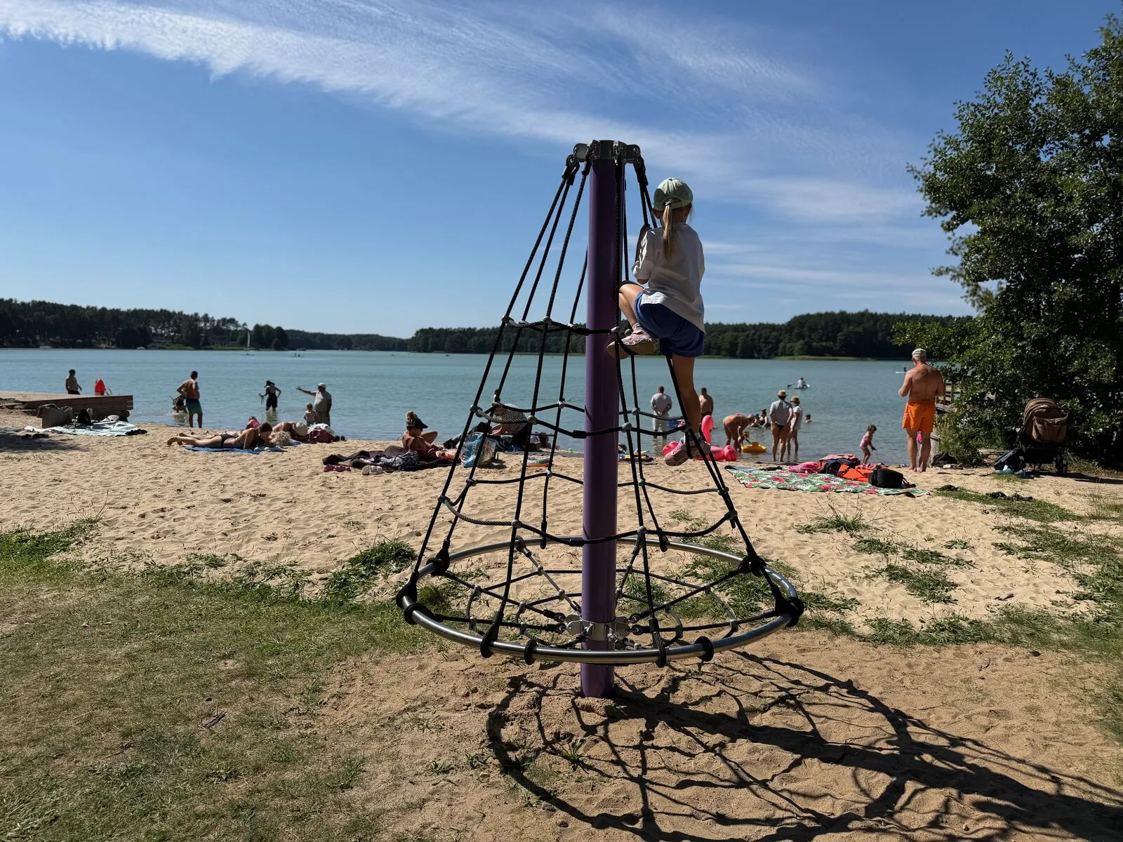 Mała piaszczysta plaża na terenie ośrodka Jasnochówka w Borsku, z siatką do wspinania dla dzieci i widokiem na Jezioro Wdzydzkie